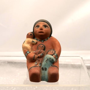 Puede incluir: Figura de cerámica que representa a una mujer sosteniendo a tres niños. La figura está pintada en tonos naranja, beige y azul, con detalles en negro. La mujer lleva un tocado negro y un collar turquesa. Los niños están en varias posiciones.
