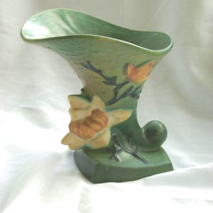 May include: A green ceramic vase with a flared, asymmetrical rim and a square base. The body features a raised, molded floral design with yellow and orange petals, dark green stems, and a small, rounded scroll-like protrusion at the bottom.