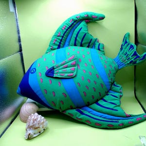 May include: A plush fish pillow in shades of green, blue, and pink. The pillow features a detailed design with stripes and spots, and is accompanied by two seashells. The fish has large fins and a tail.
