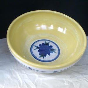 May include: A round ceramic bowl with a pale yellow interior and a white exterior. The center of the bowl features a blue floral design enclosed within a blue circular border. The exterior rim is decorated with thin blue horizontal lines and dots.