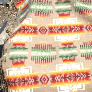 May include: A beige blanket with a geometric pattern in red, orange, yellow, green, and white. The blanket has a fringed edge. The design features repeating shapes and patterns.