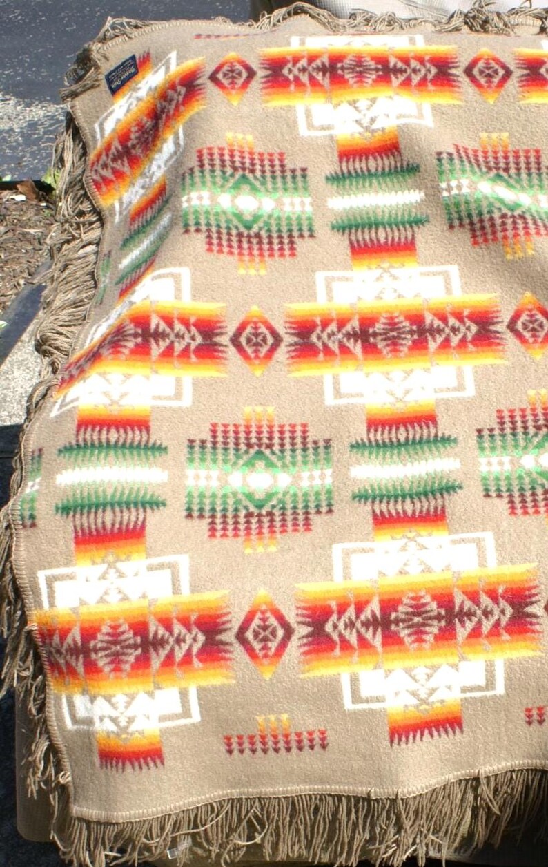 May include: A beige blanket with a geometric pattern in red, orange, yellow, green, and white. The blanket has a fringed edge. The design features repeating shapes and patterns.