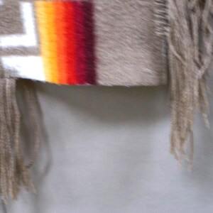 May include: Close-up of a fringed textile with a geometric pattern. The textile features a beige background with white, red, orange, and yellow stripes. The fringe is a light beige colour.