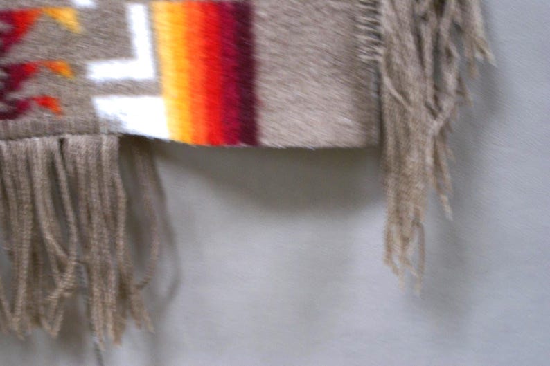 May include: Close-up of a fringed textile with a geometric pattern. The textile features a beige background with white, red, orange, and yellow stripes. The fringe is a light beige colour.