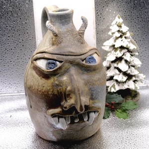 May include: A handmade ceramic jug with a face design. The jug has a detailed face with blue eyes, sharp teeth, and small horns. The colour palette includes shades of brown, grey, and blue. A small Christmas tree is in the background.