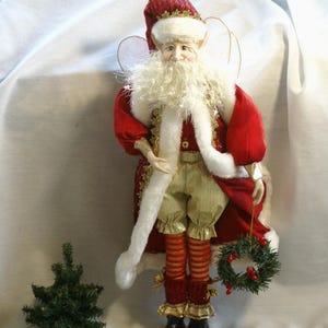 Puede incluir: Una figura decorativa de Papá Noel con un atuendo rojo y dorado, sosteniendo una pequeña corona. La figura tiene barba blanca, gorro rojo y detalles dorados. Un pequeño árbol de Navidad está al lado.