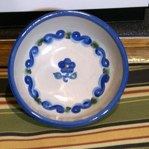 May include: A small, round ceramic bowl with a white interior and a blue rim. The bowl is decorated with blue scrollwork and a central blue flower design. The bowl is approximately 4 inches in diameter.