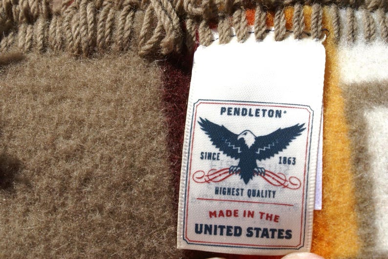 May include: Close-up of a woven textile with a tag. The tag reads "PENDLETON" with an eagle emblem and "HIGHEST QUALITY" and "MADE IN THE UNITED STATES". The textile has a brown, tan, and orange colour scheme.
