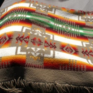 May include: A woven blanket with a Southwestern design. The blanket features a repeating pattern of geometric shapes in shades of brown, white, red, orange, and green. The blanket has a fringe along the bottom edge.