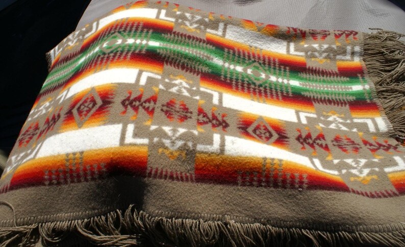 May include: A woven blanket with a Southwestern design. The blanket features a repeating pattern of geometric shapes in shades of brown, white, red, orange, and green. The blanket has a fringe along the bottom edge.