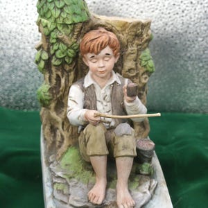 Puede incluir: Una figura de cerámica de una persona joven con cabello castaño rojizo, vistiendo camisa blanca, chaleco marrón y pantalones beige, sentada descalza contra un tronco de árbol. La figura sostiene una pequeña pipa y un palo fino. La base tiene la etiqueta HUCK FINN.