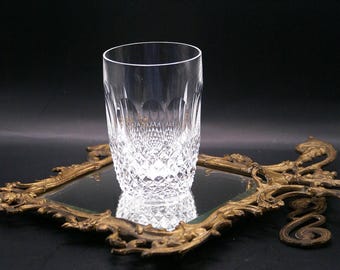May include: A clear, cut-crystal tumbler glass with a diamond pattern, standing on a decorative, antique gold-colored mirror. The glass is cylindrical with a smooth rim. The mirror has an ornate, vintage frame.