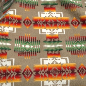May include: A tan blanket with a Southwestern geometric pattern. The design features repeating shapes in white, red, orange, and green. The blanket has a soft, textured appearance, with a fringe detail visible on the edge.