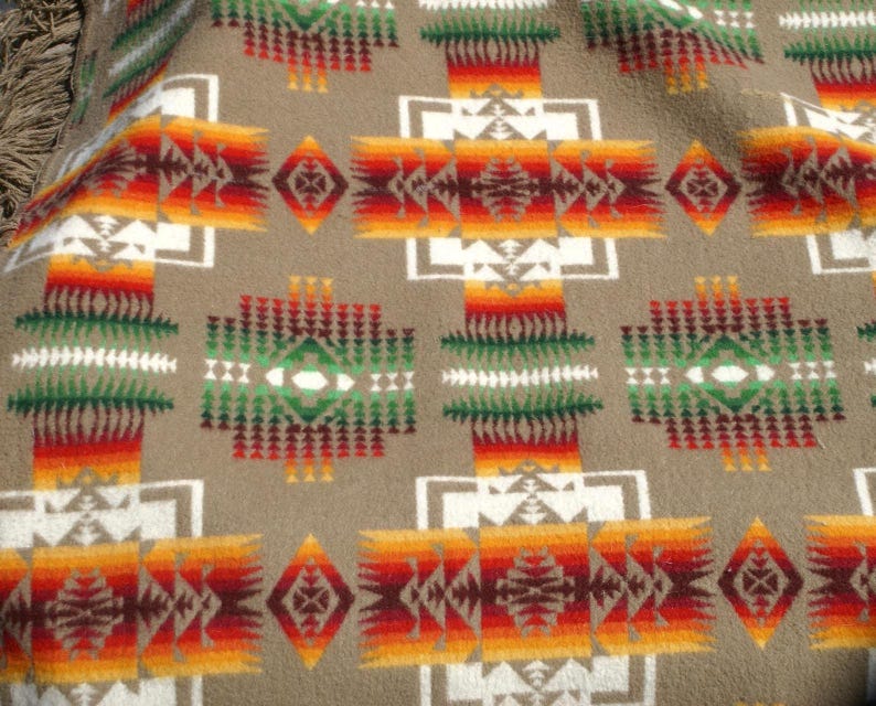 May include: A tan blanket with a Southwestern geometric pattern. The design features repeating shapes in white, red, orange, and green. The blanket has a soft, textured appearance, with a fringe detail visible on the edge.