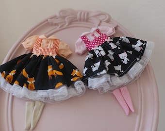 May include: Two doll dresses with white lace trim. One dress is black with orange fox print and a red and white gingham bodice. The other dress is black with white cat print and a pink polka dot bodice.