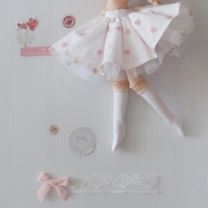 Op de afbeelding: Een witte poppenrok met roze hartjes en een witte tule overlay. De rok wordt gedragen door een pop met witte sokken en een roze strik.