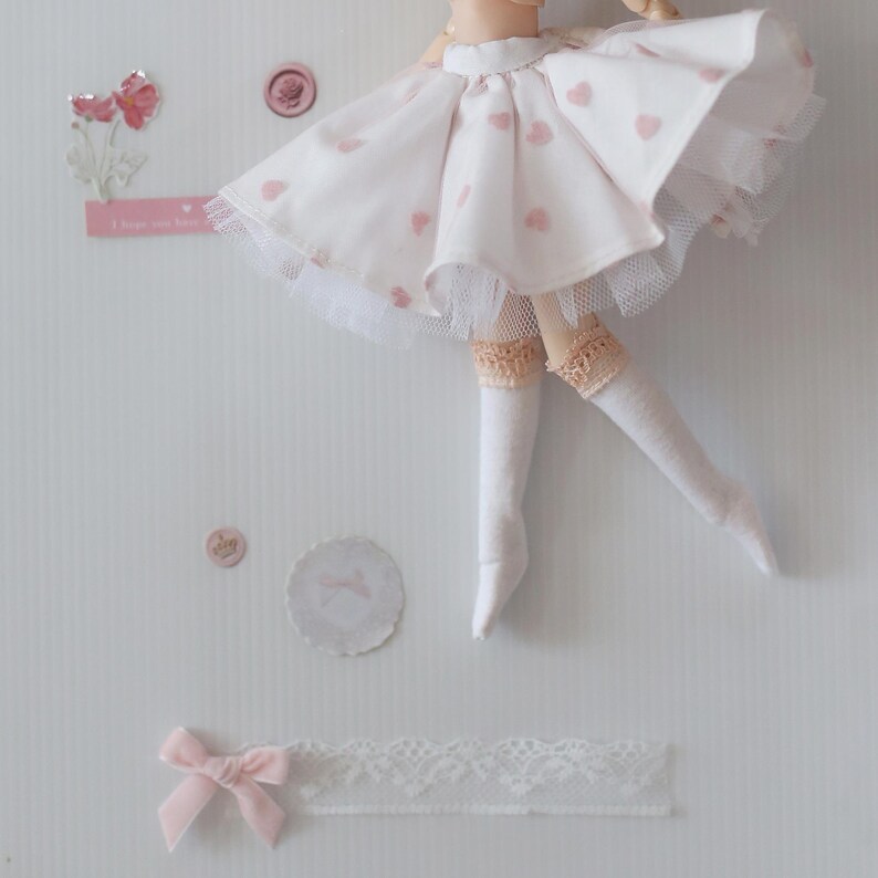 Op de afbeelding: Een witte poppenrok met roze hartjes en een witte tule overlay. De rok wordt gedragen door een pop met witte sokken en een roze strik.