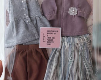 May include: A set of doll clothing including a gray sleeveless vest with white buttons, a mauve long-sleeved jacket with a white floral accent, brown trousers, and a blue and white striped skirt. A square sticker reads: YOUR PURCHASE MADE MY DAY & I HOPE THIS PACKAGE BRIGHTENS YOURS.