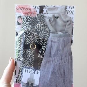 Puede incluir: Un conjunto de ropa de mu&ntilde;eca en una bolsa de pl&aacute;stico transparente. El conjunto incluye un abrigo estampado en blanco y negro, un su&eacute;ter gris de cuello alto con el logotipo de Prada, una falda de tul gris y una revista Vogue en miniatura.