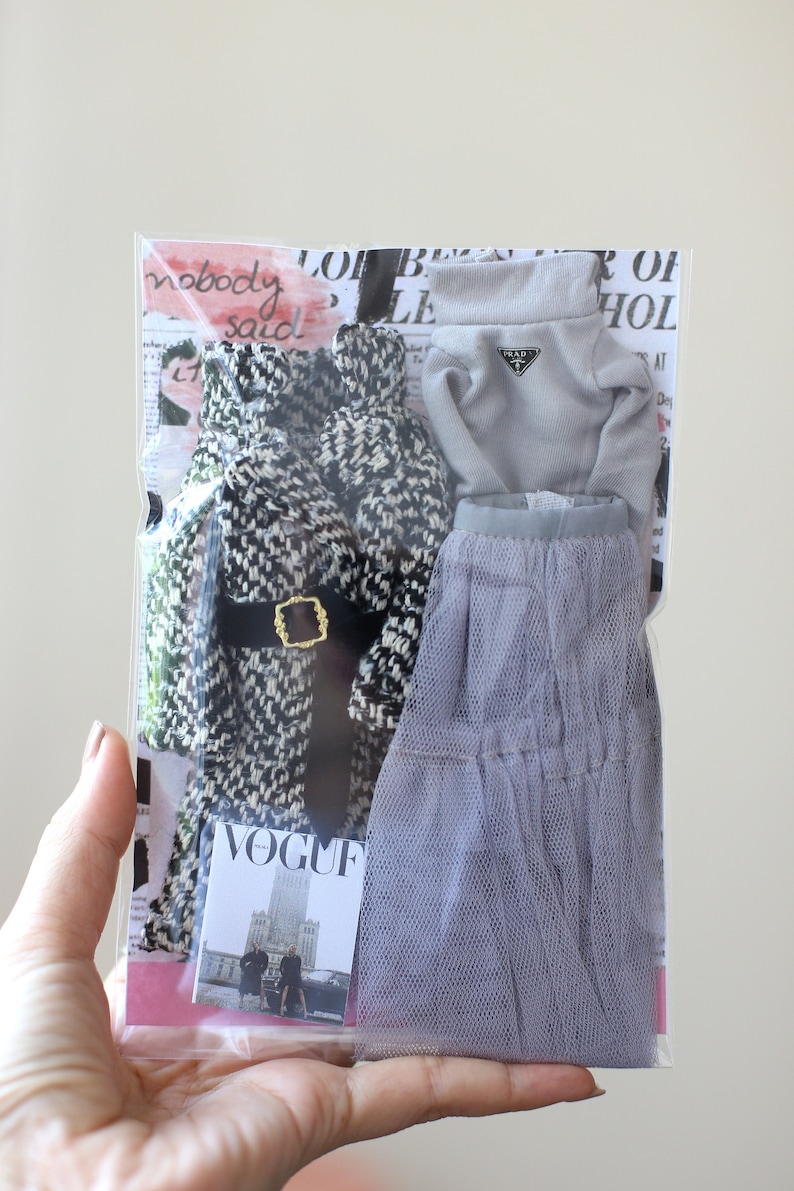 Puede incluir: Un conjunto de ropa de mu&ntilde;eca en una bolsa de pl&aacute;stico transparente. El conjunto incluye un abrigo estampado en blanco y negro, un su&eacute;ter gris de cuello alto con el logotipo de Prada, una falda de tul gris y una revista Vogue en miniatura.