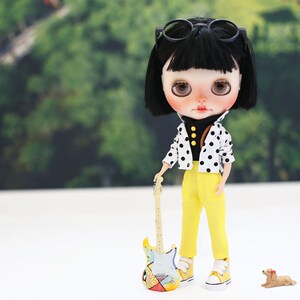 May include: A doll with black hair and glasses wearing a black and white polka dot shirt and yellow pants, holding a yellow and blue toy guitar.
