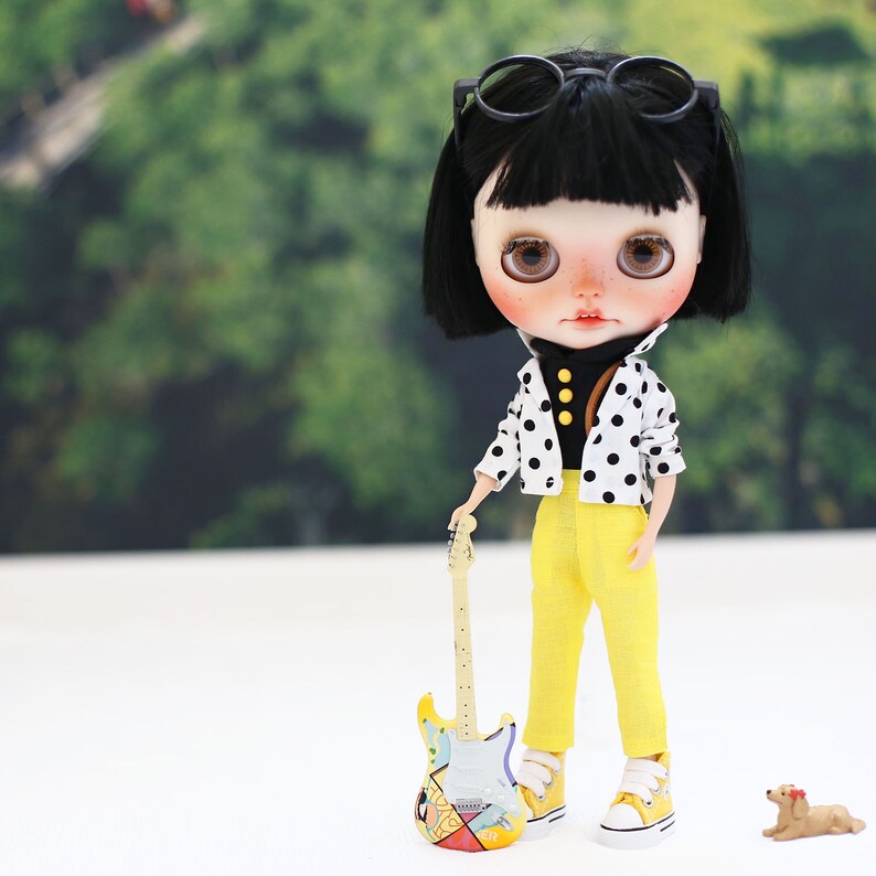 May include: A doll with black hair and glasses wearing a black and white polka dot shirt and yellow pants, holding a yellow and blue toy guitar.