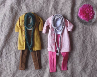 May include: Two doll outfits: one mustard yellow coat with brown pants and a green braided scarf, and a pink cardigan with pink pants and a gray braided scarf. A pink flower in a glass is in the background.