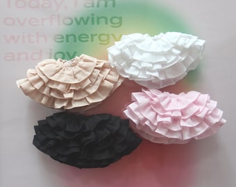 May include: Four ruffled fabric skirts in different colors: black, white, pink, and beige. The skirts are layered with multiple ruffles and are arranged on a white surface.