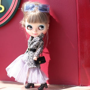 Puede incluir: Una mu&ntilde;eca con cabello rubio que lleva un abrigo estampado en blanco y negro, una falda de tul blanca y gafas de sol azules. La mu&ntilde;eca lleva un bolso negro y est&aacute; de pie frente a una pared roja.