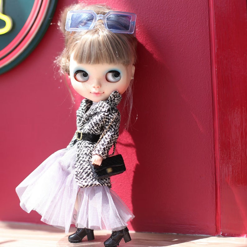 Puede incluir: Una mu&ntilde;eca con cabello rubio que lleva un abrigo estampado en blanco y negro, una falda de tul blanca y gafas de sol azules. La mu&ntilde;eca lleva un bolso negro y est&aacute; de pie frente a una pared roja.