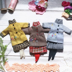 May include: Three miniature doll outfits with embroidered floral details. The outfits are a yellow coat with a black and white houndstooth skirt, a brown coat with a red plaid skirt, and a blue coat with a black and white plaid skirt.