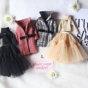 Puede incluir: Cuatro conjuntos de mu&ntilde;ecas en miniatura, que incluyen un vestido negro con falda de tul, una chaqueta de tweed roja y blanca, una chaqueta a cuadros y un su&eacute;ter beige con falda de tul. Los conjuntos est&aacute;n dispuestos sobre una superficie blanca con un peque&ntilde;o cartel que dice "&iexcl;Recuerda hacer tu pedido!"