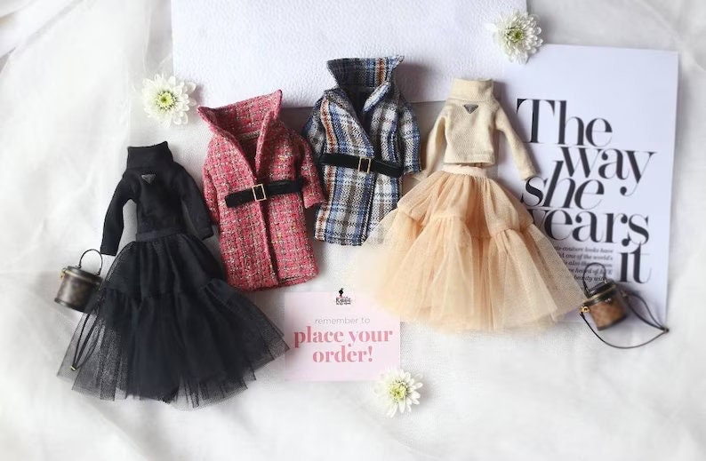 Puede incluir: Cuatro conjuntos de mu&ntilde;ecas en miniatura, que incluyen un vestido negro con falda de tul, una chaqueta de tweed roja y blanca, una chaqueta a cuadros y un su&eacute;ter beige con falda de tul. Los conjuntos est&aacute;n dispuestos sobre una superficie blanca con un peque&ntilde;o cartel que dice "&iexcl;Recuerda hacer tu pedido!"