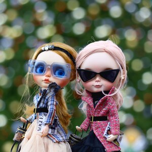 Puede incluir: Dos mu&ntilde;ecas de moda con gafas de sol y atuendos elegantes. Una mu&ntilde;eca tiene el pelo rubio y lleva una chaqueta de cuadros azules, una falda de tul y un bolso negro. La otra mu&ntilde;eca tiene el pelo rosa y lleva una chaqueta de tweed rosa, una falda de tul negra y un bolso negro. Ambas mu&ntilde;ecas llevan peque&ntilde;os bolsos con la palabra "Vogue" impresa.
