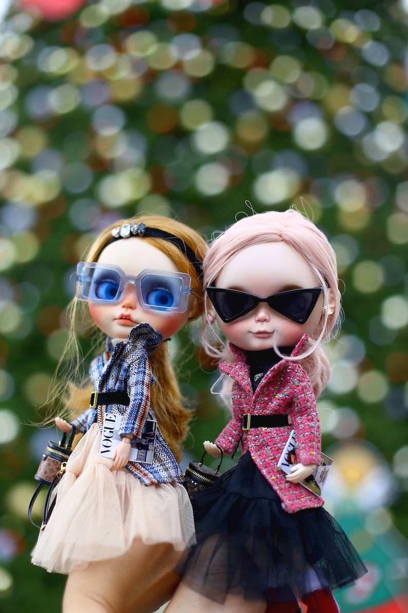 Puede incluir: Dos mu&ntilde;ecas de moda con gafas de sol y atuendos elegantes. Una mu&ntilde;eca tiene el pelo rubio y lleva una chaqueta de cuadros azules, una falda de tul y un bolso negro. La otra mu&ntilde;eca tiene el pelo rosa y lleva una chaqueta de tweed rosa, una falda de tul negra y un bolso negro. Ambas mu&ntilde;ecas llevan peque&ntilde;os bolsos con la palabra "Vogue" impresa.