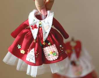 May include: A red dress with white trim and a white collar. The dress has a white pocket with a red and white checkered pattern and a small embroidered Christmas tree on the front. The dress has a white tulle underskirt.