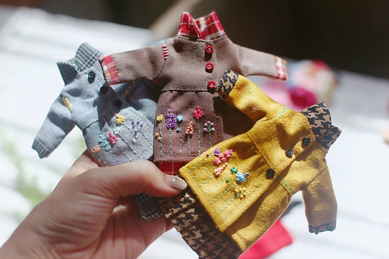 May include: Four miniature doll clothes with embroidered floral designs. The clothes are in shades of brown, grey, yellow, and red.