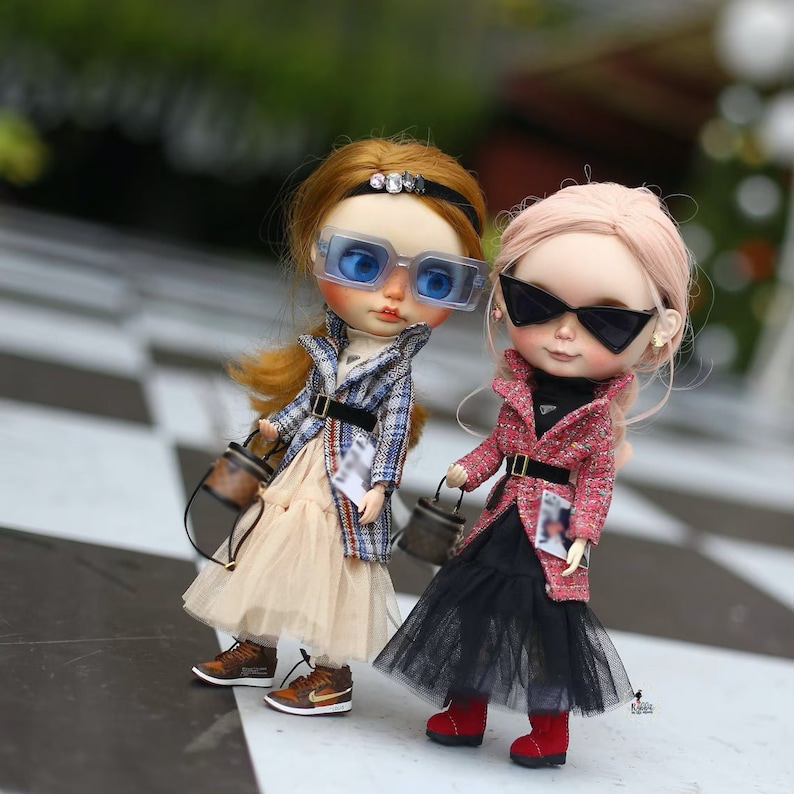 Puede incluir: Dos mu&ntilde;ecas con atuendos elegantes y gafas de sol. Una mu&ntilde;eca lleva una chaqueta de cuadros y una falda de tul, mientras que la otra lleva una chaqueta roja y una falda de tul negra. Ambas mu&ntilde;ecas llevan bolsos peque&ntilde;os.