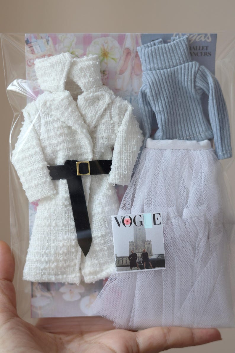 Puede incluir: Un abrigo blanco texturizado con un cintur&oacute;n negro, un su&eacute;ter azul claro de cuello alto y una falda de tul blanca. Una revista con la palabra "VOGUE" en la portada es visible en la imagen.