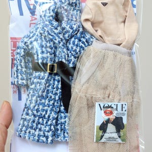 Puede incluir: Un conjunto de prendas de vestir en una bolsa de pl&aacute;stico transparente. El conjunto incluye una chaqueta de tweed azul y blanco, un jersey de cuello alto beige, una falda de tul beige y una revista Vogue con Harry Styles.