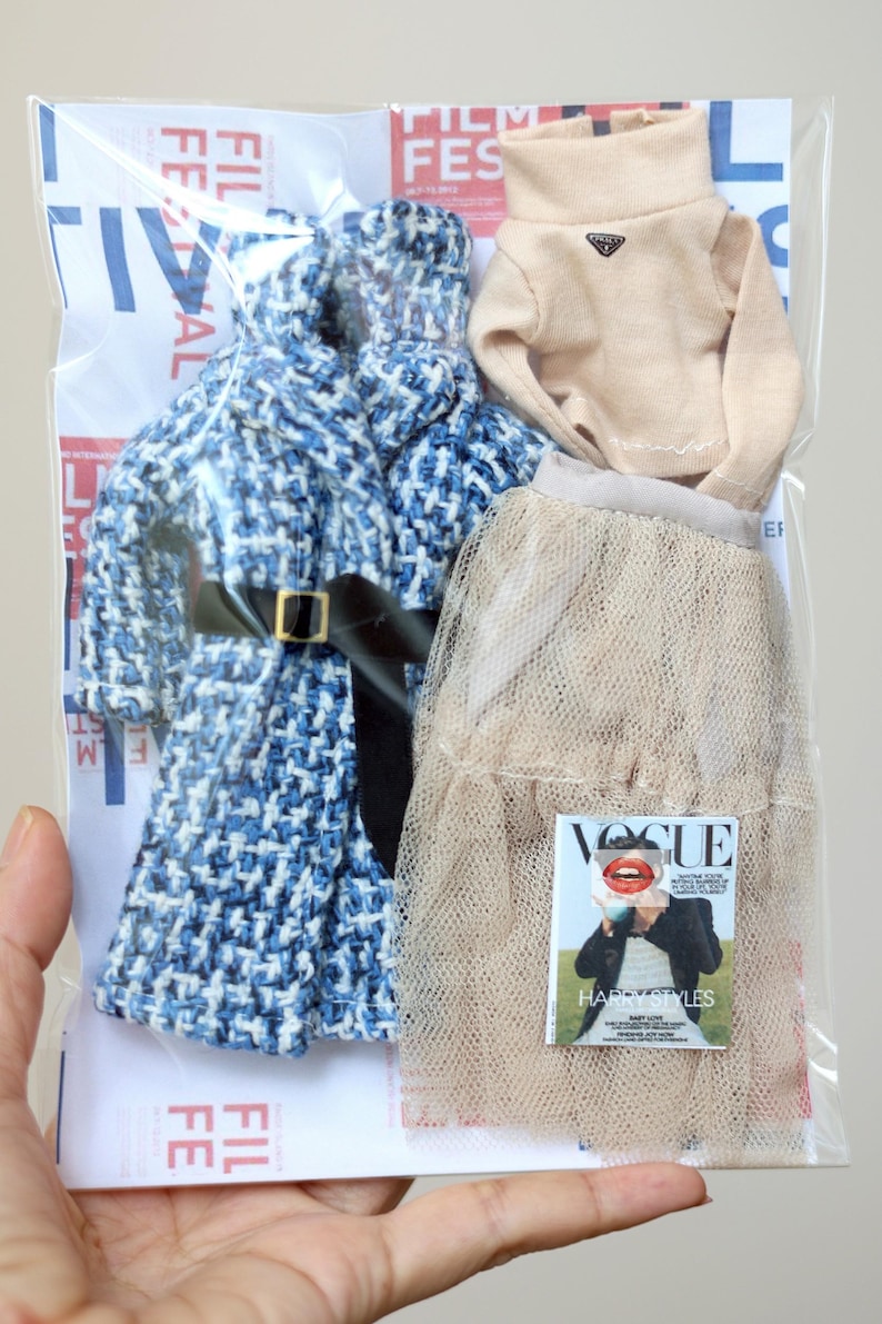 Puede incluir: Un conjunto de prendas de vestir en una bolsa de pl&aacute;stico transparente. El conjunto incluye una chaqueta de tweed azul y blanco, un jersey de cuello alto beige, una falda de tul beige y una revista Vogue con Harry Styles.