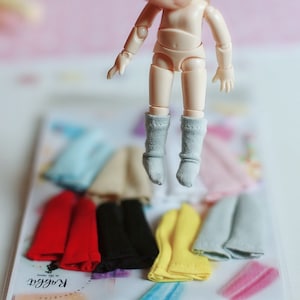 May include: A small doll wearing a pair of gray knee-high socks. The doll is floating above a variety of colorful clothing items, including red, yellow, black, and blue.