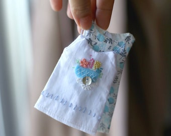May include: A white sleeveless dress with a blue and white floral print. The dress has a blue embroidered flower design on the front with a white button in the center.