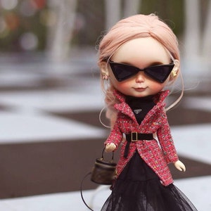 Puede incluir: Una mu&ntilde;eca con el pelo rosa que lleva una chaqueta de tweed roja y negra, una falda negra, medias negras y zapatos rojos. La mu&ntilde;eca lleva un peque&ntilde;o bolso marr&oacute;n y gafas de sol negras.