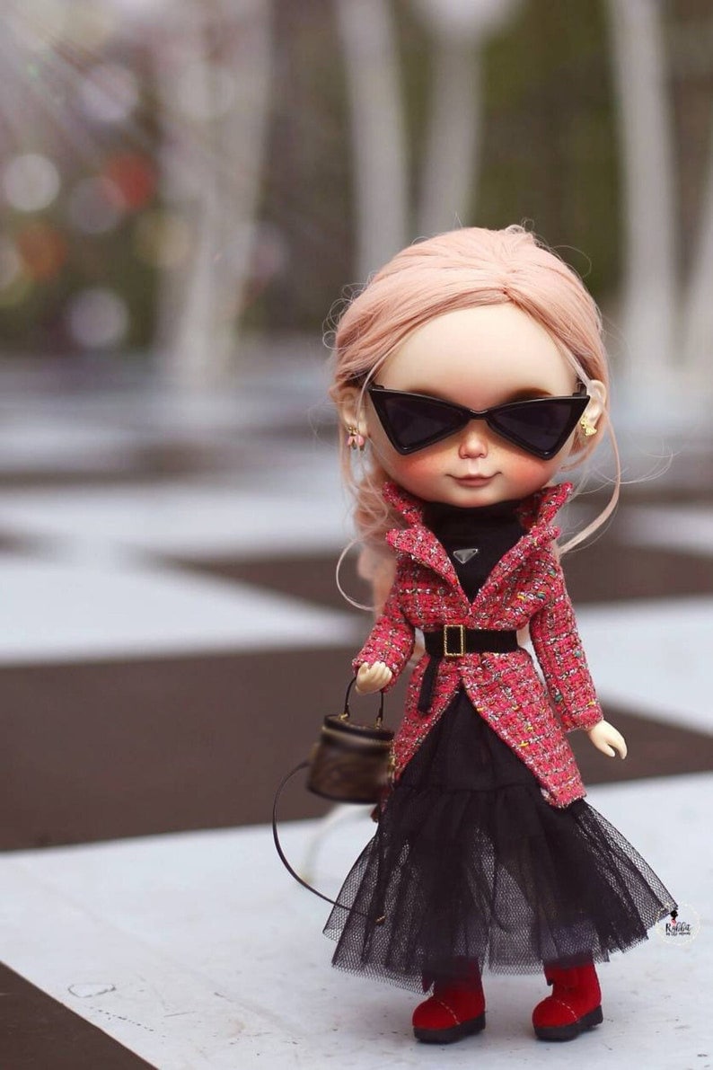 Puede incluir: Una mu&ntilde;eca con el pelo rosa que lleva una chaqueta de tweed roja y negra, una falda negra, medias negras y zapatos rojos. La mu&ntilde;eca lleva un peque&ntilde;o bolso marr&oacute;n y gafas de sol negras.