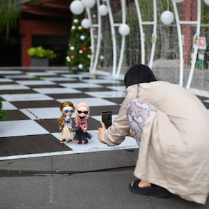 Puede incluir: Una persona tomando una foto de tres mu&ntilde;ecas con gafas de sol y atuendos elegantes. Las mu&ntilde;ecas est&aacute;n de pie sobre un suelo de ajedrez blanco y negro frente a un &aacute;rbol de Navidad.