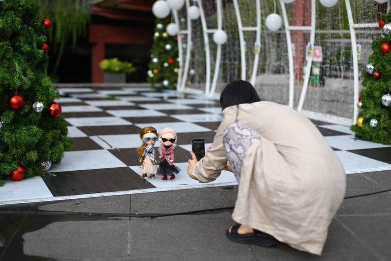 Puede incluir: Una persona tomando una foto de tres mu&ntilde;ecas con gafas de sol y atuendos elegantes. Las mu&ntilde;ecas est&aacute;n de pie sobre un suelo de ajedrez blanco y negro frente a un &aacute;rbol de Navidad.