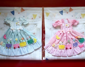 Könnte beinhalten: Zwei Puppenkleider, eines hellblau und eines rosa, jeweils mit einzigartigem Design. Das blaue Kleid hat ein Blumen- und Schmetterlingsthema, während das rosa Kleid Punkte hat. Beide Kleider haben Rüschenröcke und dekorative Akzente.