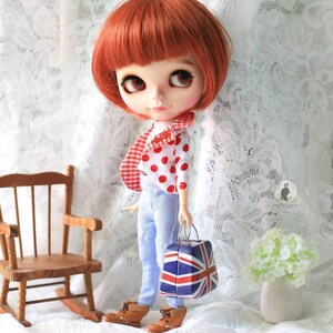 May include: A red and white polka dot shirt with a red and white checkered pattern on the back, worn by a doll with red hair and blue jeans. The doll is holding a small suitcase with a Union Jack design.