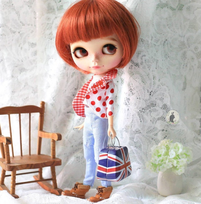 May include: A red and white polka dot shirt with a red and white checkered pattern on the back, worn by a doll with red hair and blue jeans. The doll is holding a small suitcase with a Union Jack design.